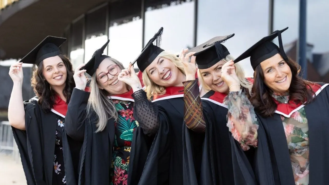 Five people in graduation gowns and mortarboards smile together outside a building.