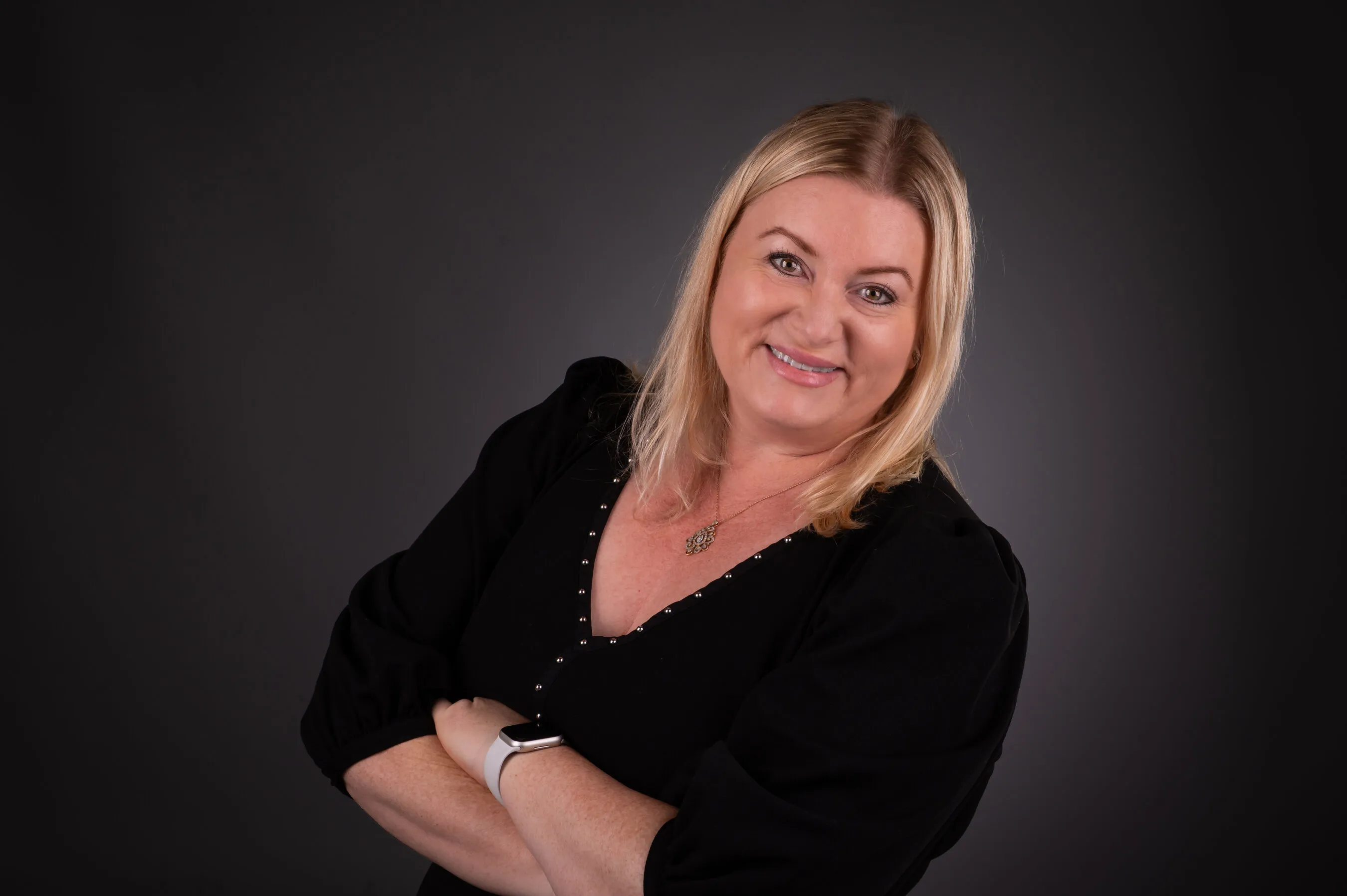 A smiling blonde woman in a black top poses with her arms crossed against a dark background.