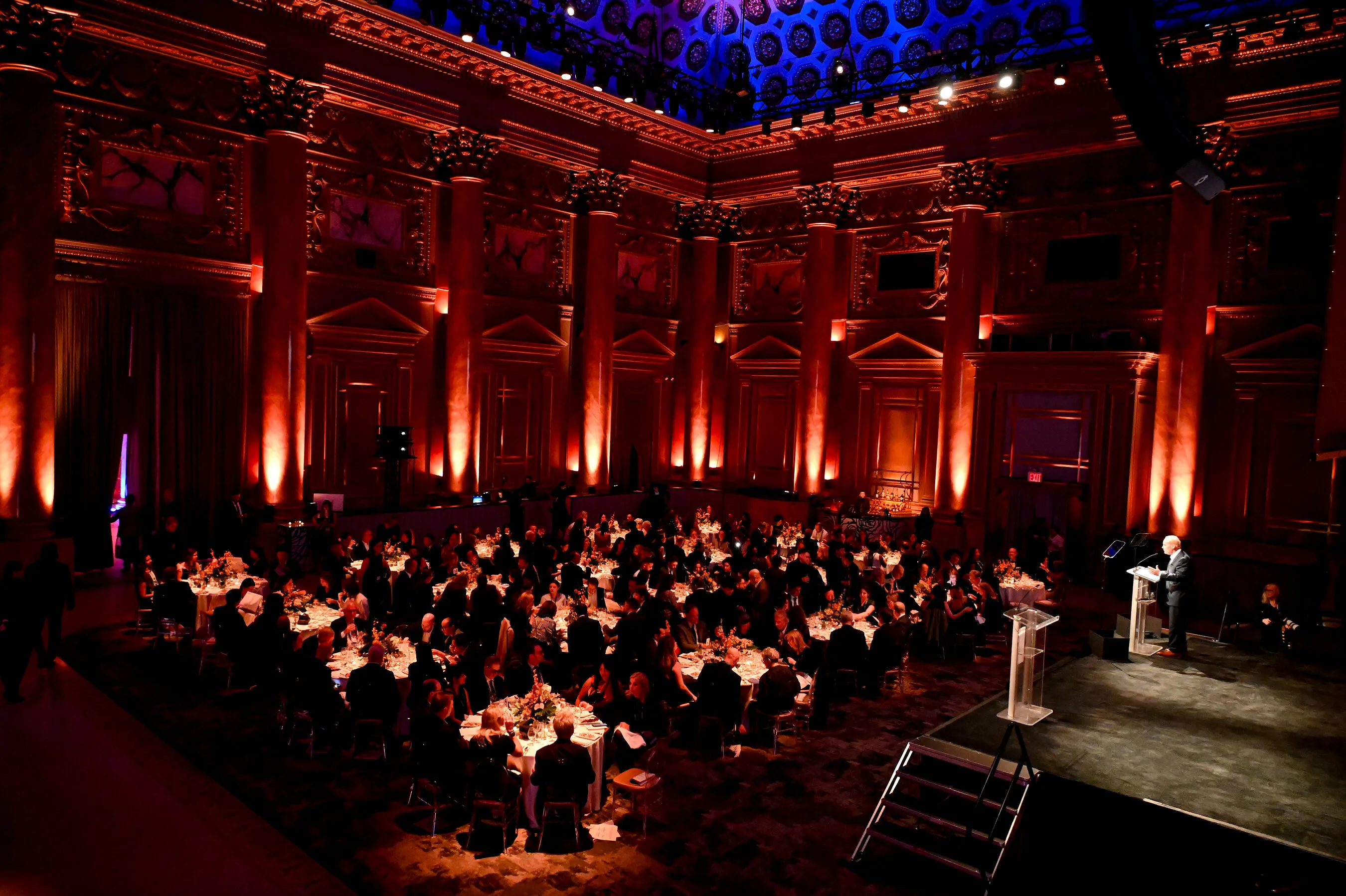 Guests seated at banquet tables in an ornate hall lit red, with a speaker at a podium.