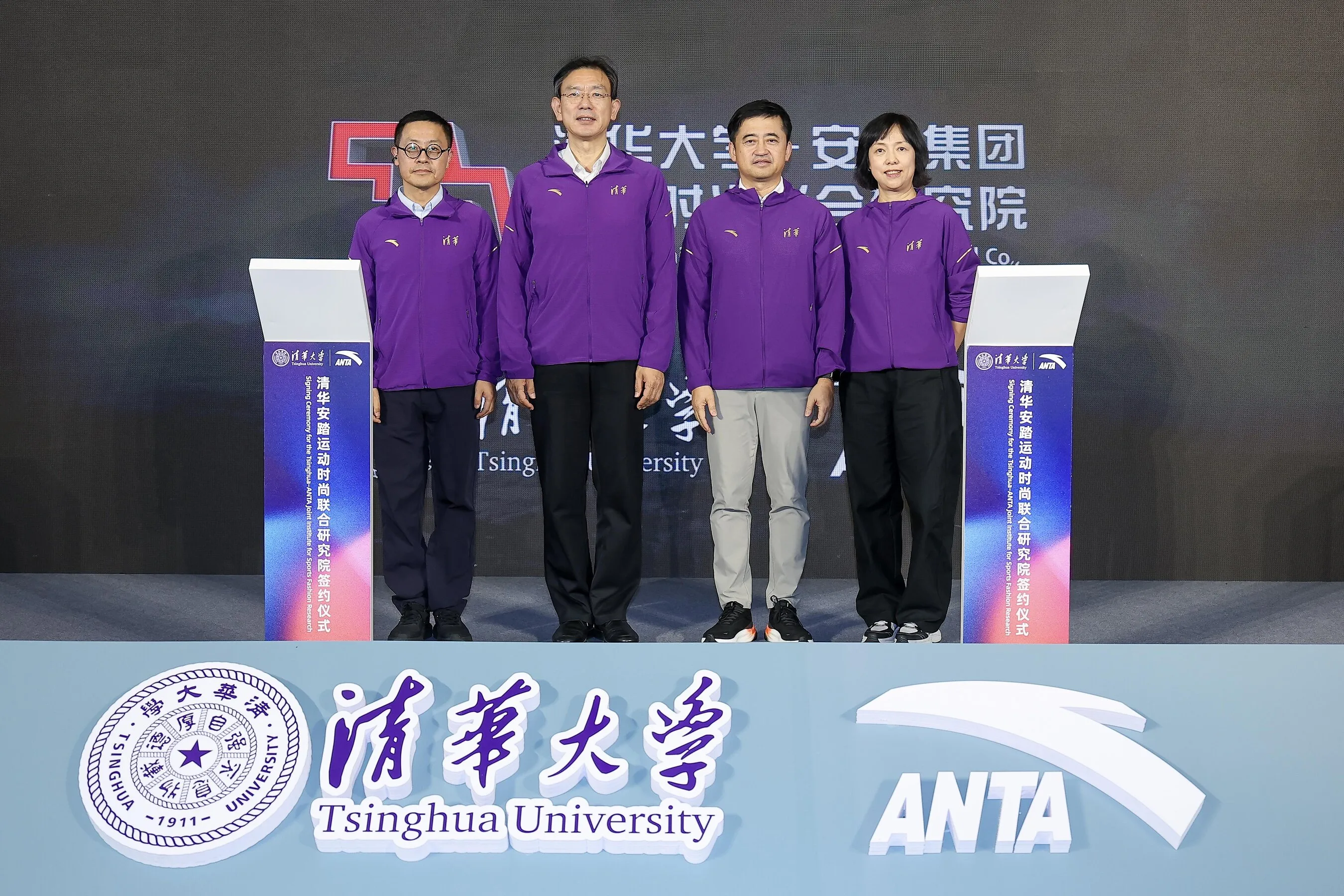 Four people in purple jackets stand between podiums at a Tsinghua University and Anta signing event.