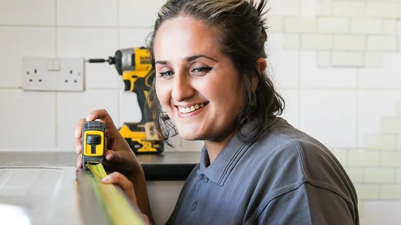 A smiling apprentice joiner measures a worktop with a tape measure in a tiled kitchen.