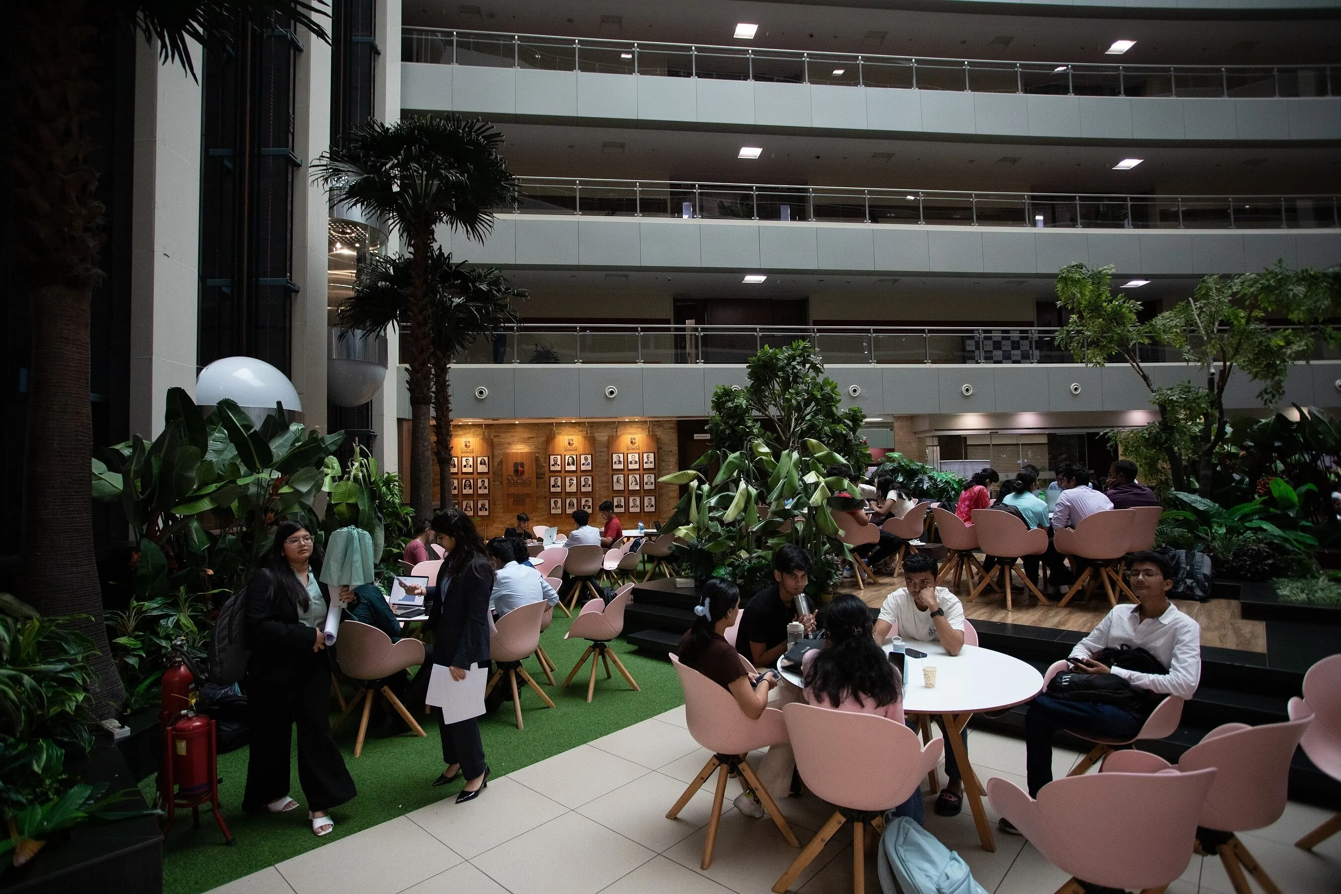 Students seated in a plant-filled indoor atrium with pink chairs and tables.
