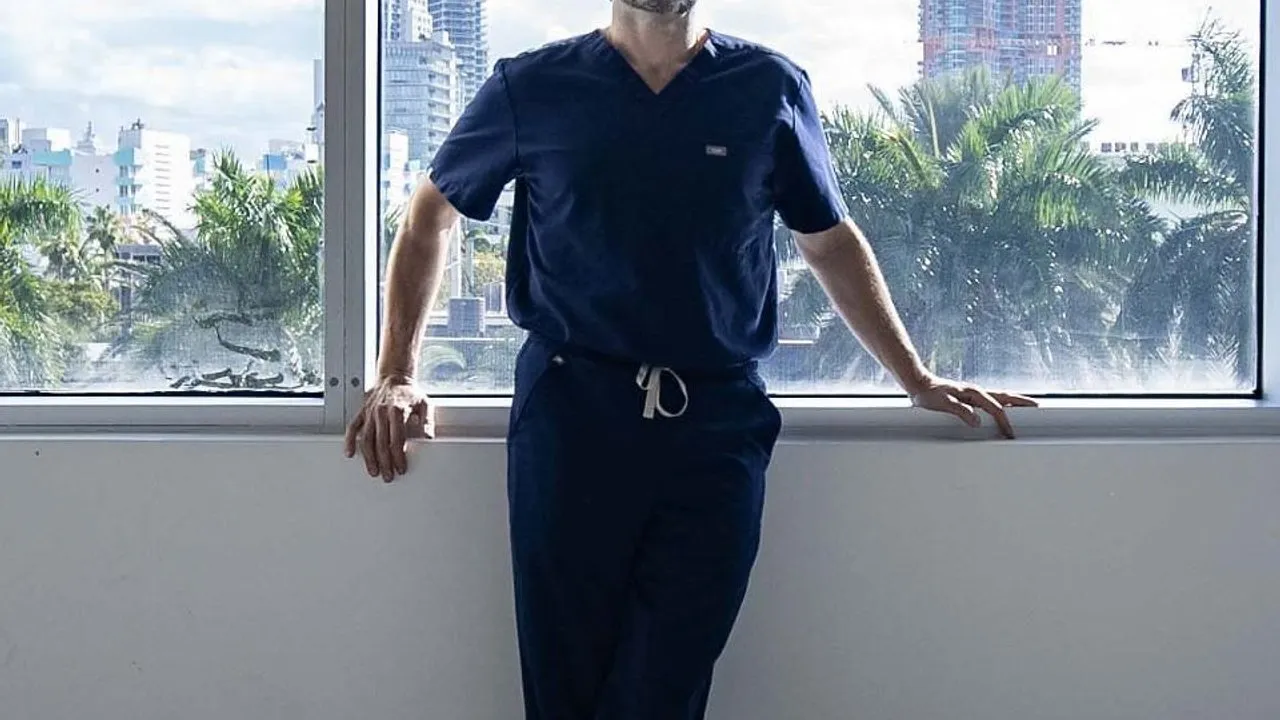 A man in navy medical scrubs stands by a window overlooking palm trees and high-rise buildings.
