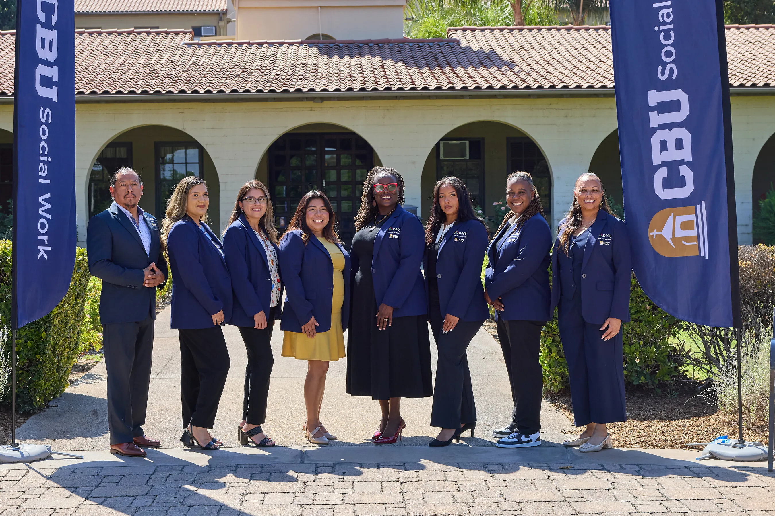 Eight people in navy blazers stand between CBU Social Work banners outside a building.
