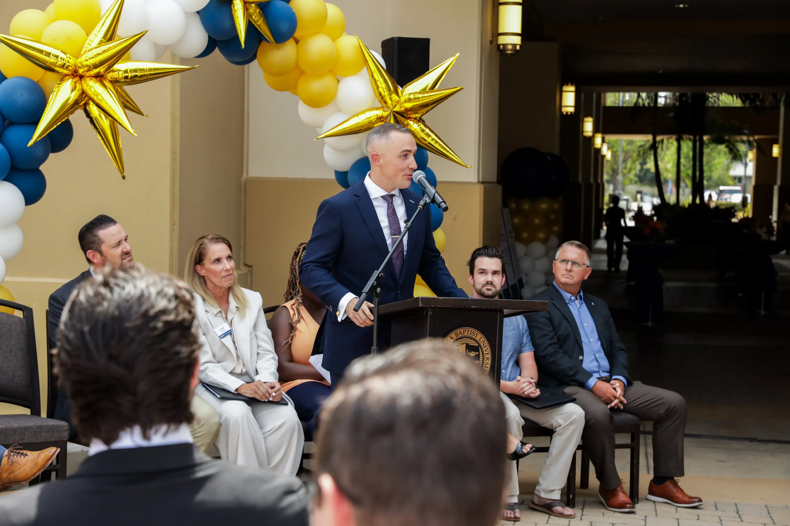 A man speaks at a podium beside seated guests under blue, white and gold balloons.
