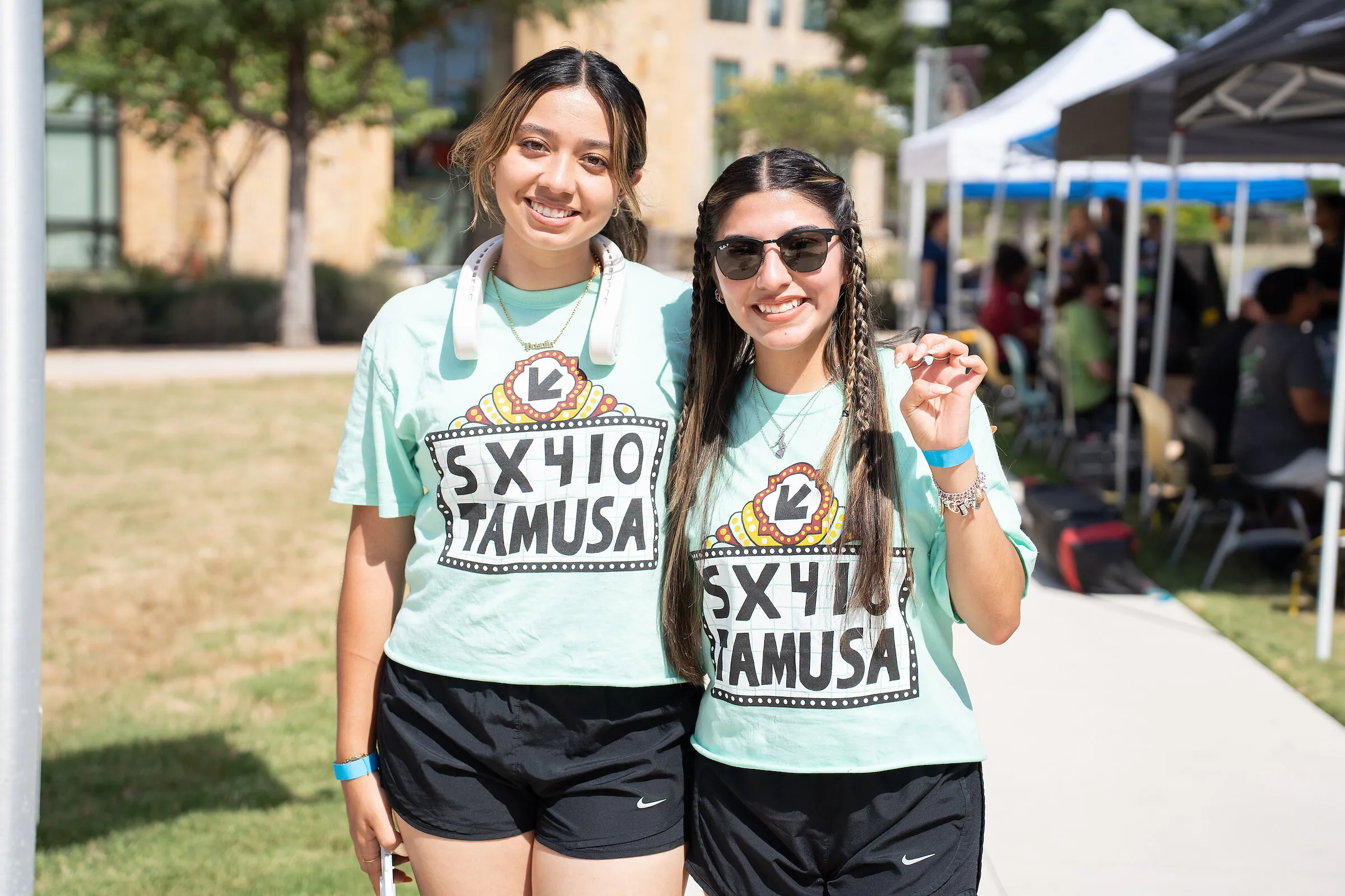 Two women wearing turquoise SX410 TAMUSA T-shirts at an outdoor event.