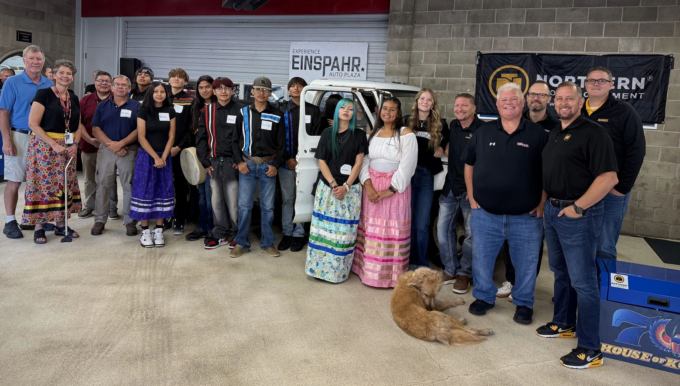 A group of people stands around a white vehicle in a garage, with a dog lying in front.