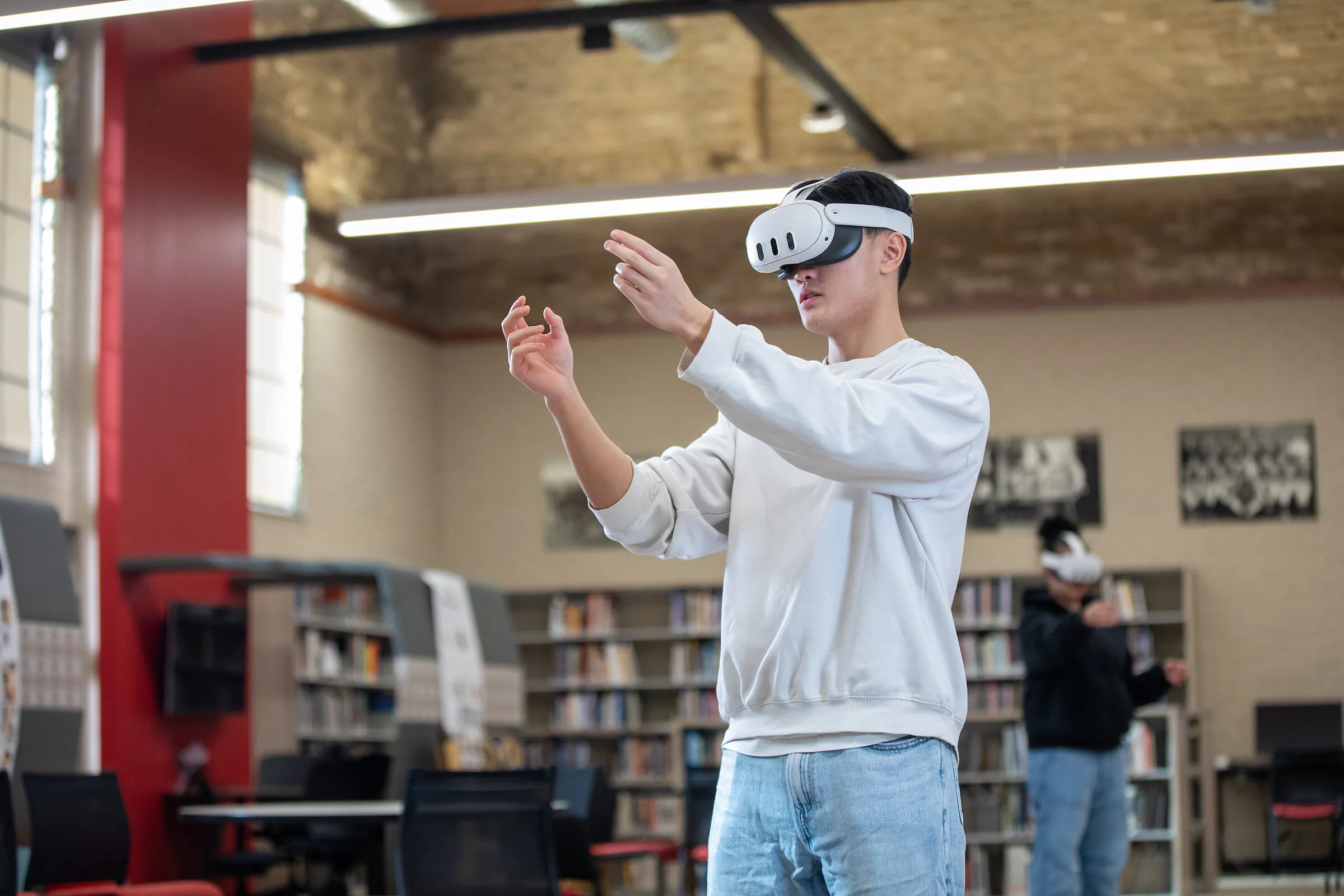 A person wearing a VR headset gestures in a library, with another headset user behind.