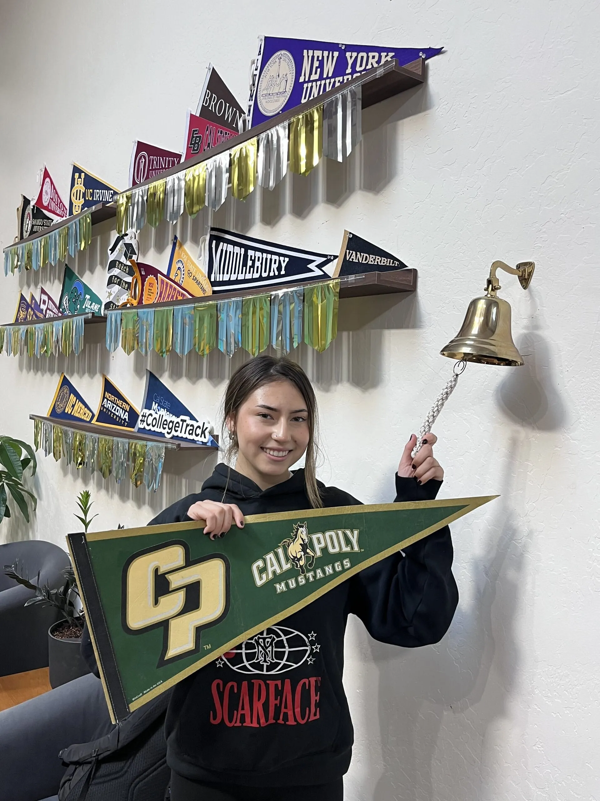A woman holds a Cal Poly pennant and rings a wall-mounted bell.