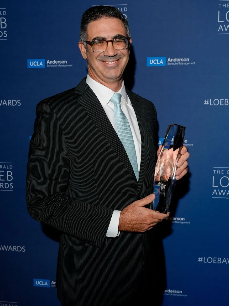Marcelo Prince in a pinstripe suit holding an award against a UCLA Anderson backdrop.