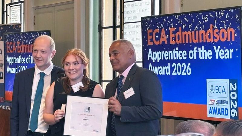 Remi Newman holds an Apprentice of the Year certificate between two people at an awards event.