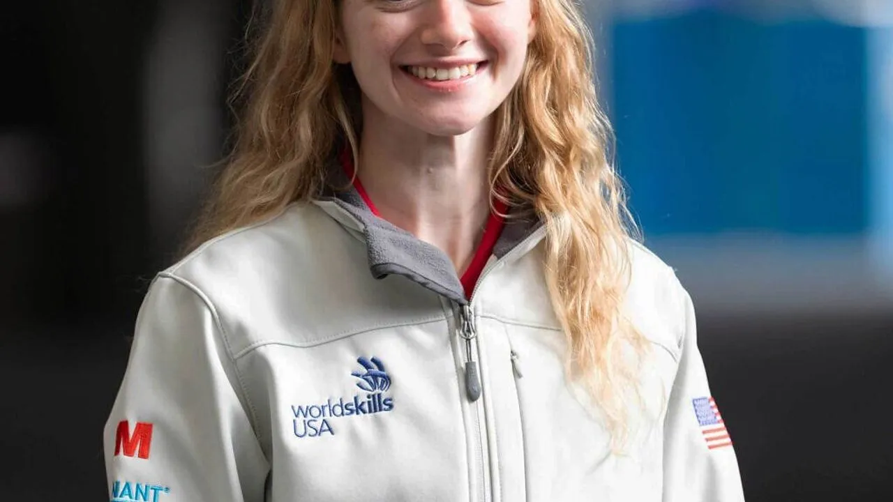 A smiling woman wears a white WorldSkills USA jacket with an American flag patch.
