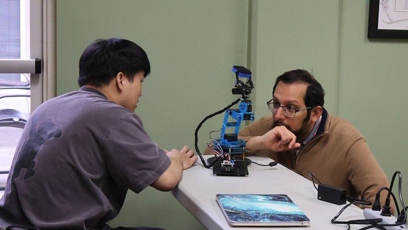Two men examine a small blue robotic arm on a table.