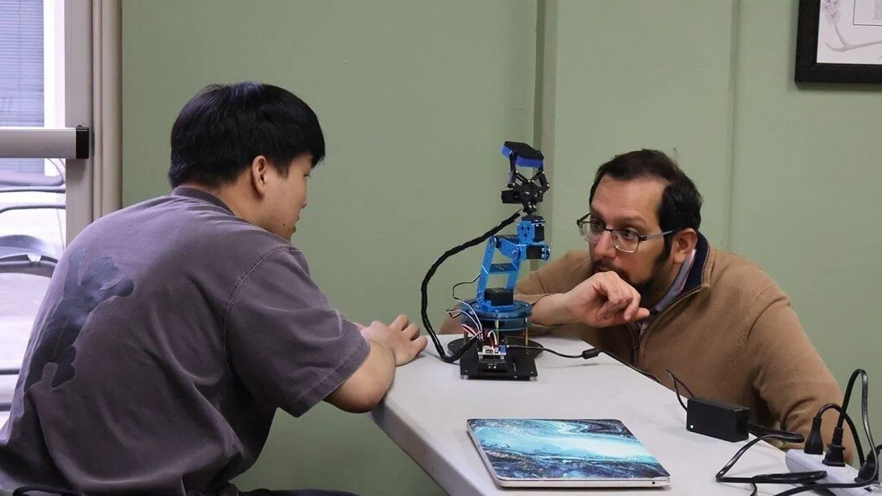 Two men examine a small blue robotic arm on a table.
