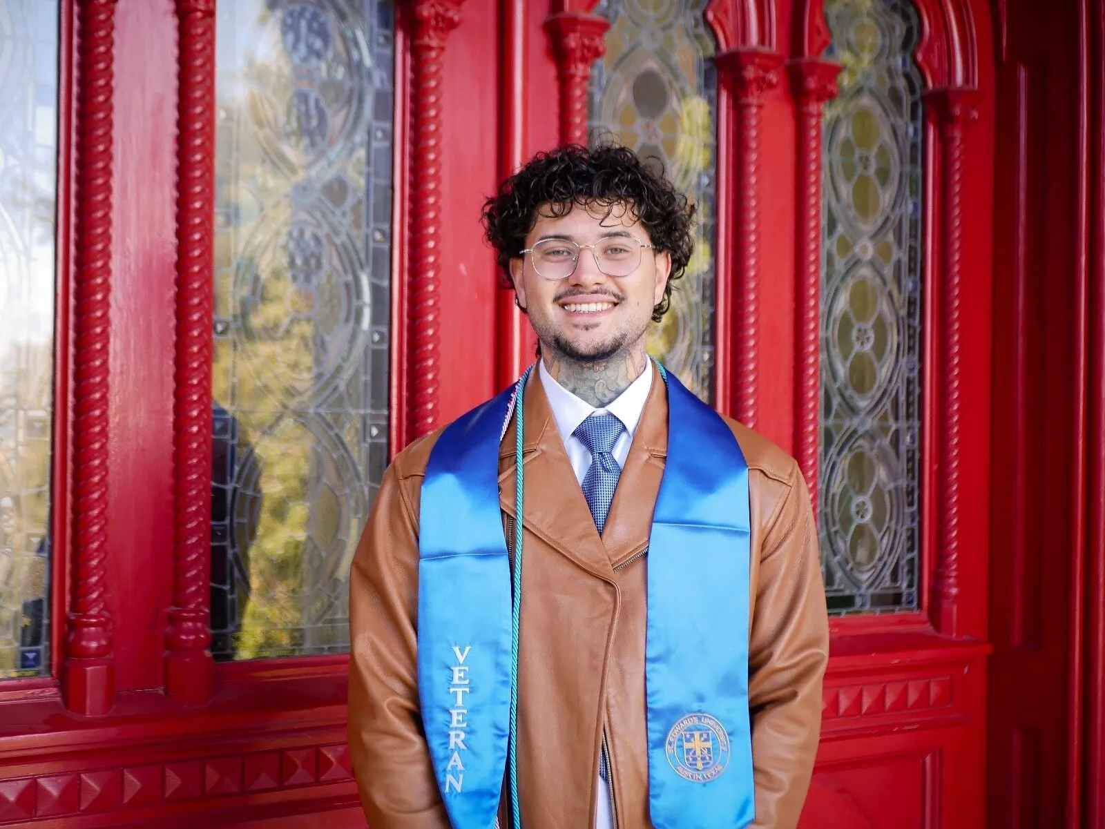 A smiling man in glasses, tie and blue graduation stole stands before ornate red doors.