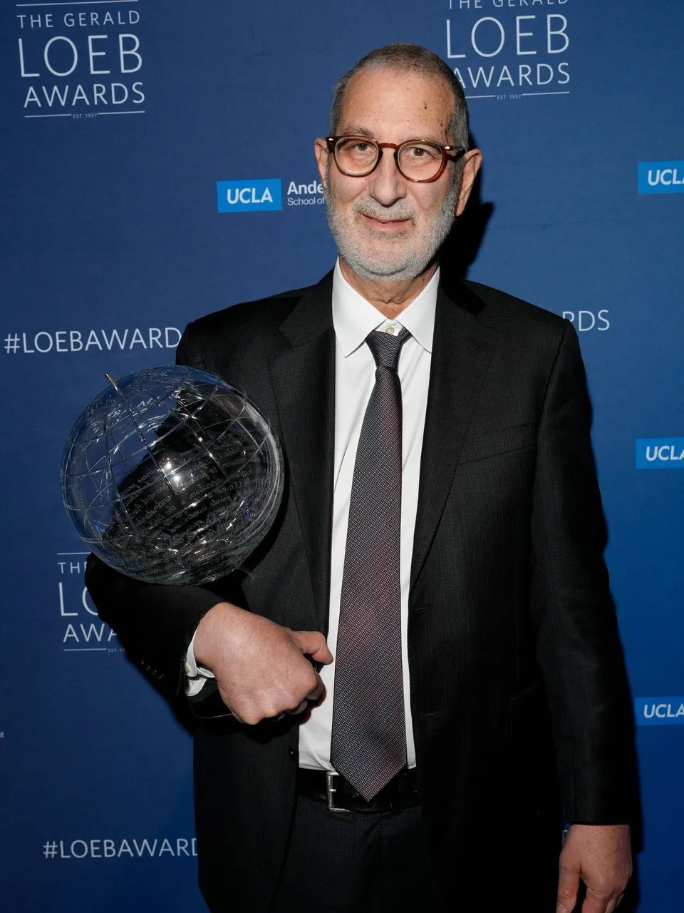 Stephen Engelberg holds a glass globe award in front of a blue Loeb Awards backdrop.