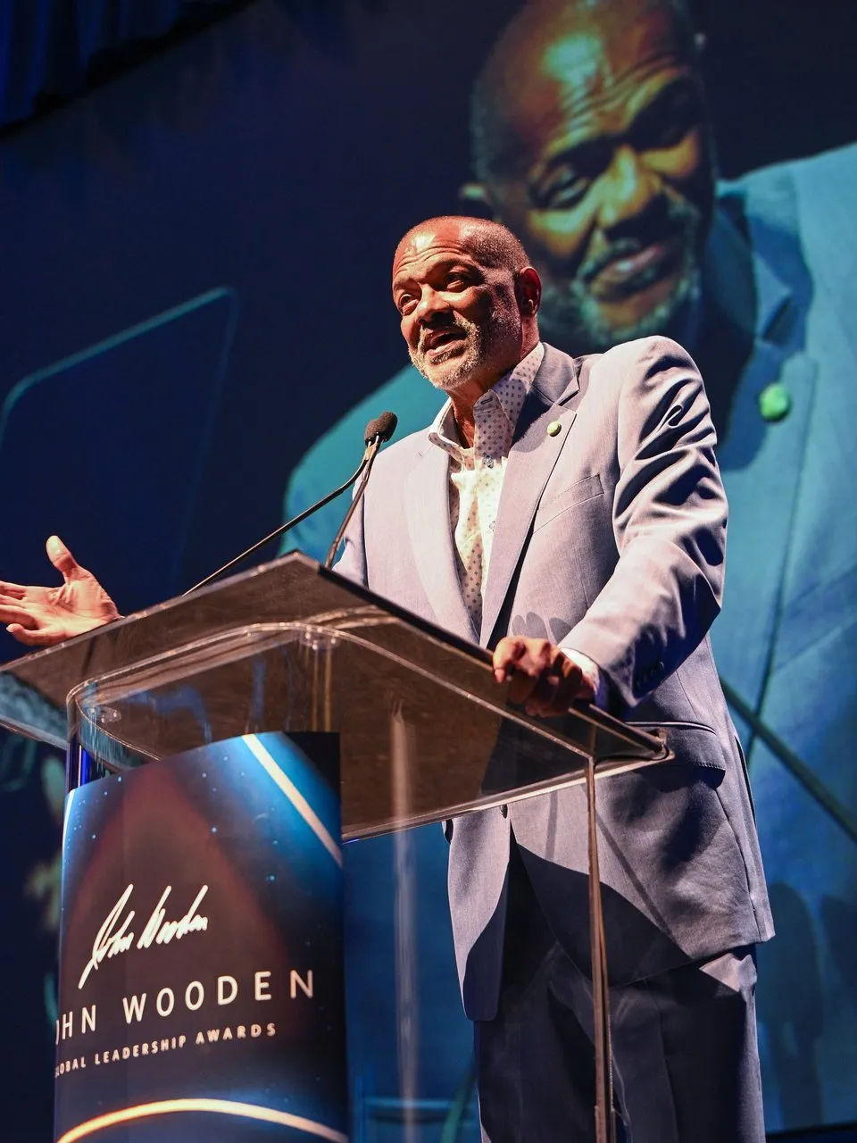 Marques Johnson speaks at a podium with his image projected on a large screen behind him.