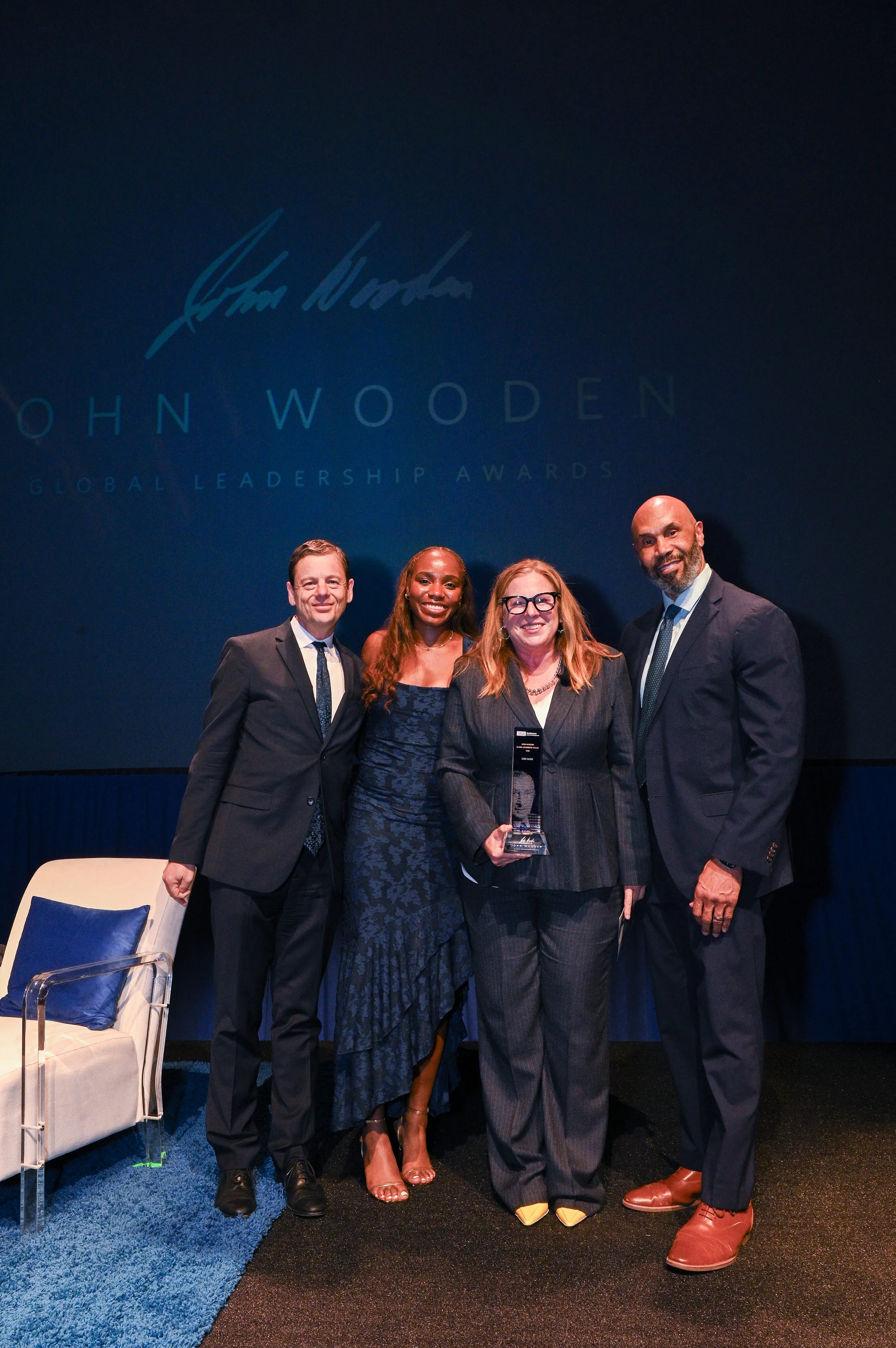 Four people pose with an award on a stage beneath a John Wooden Global Leadership Awards backdrop.