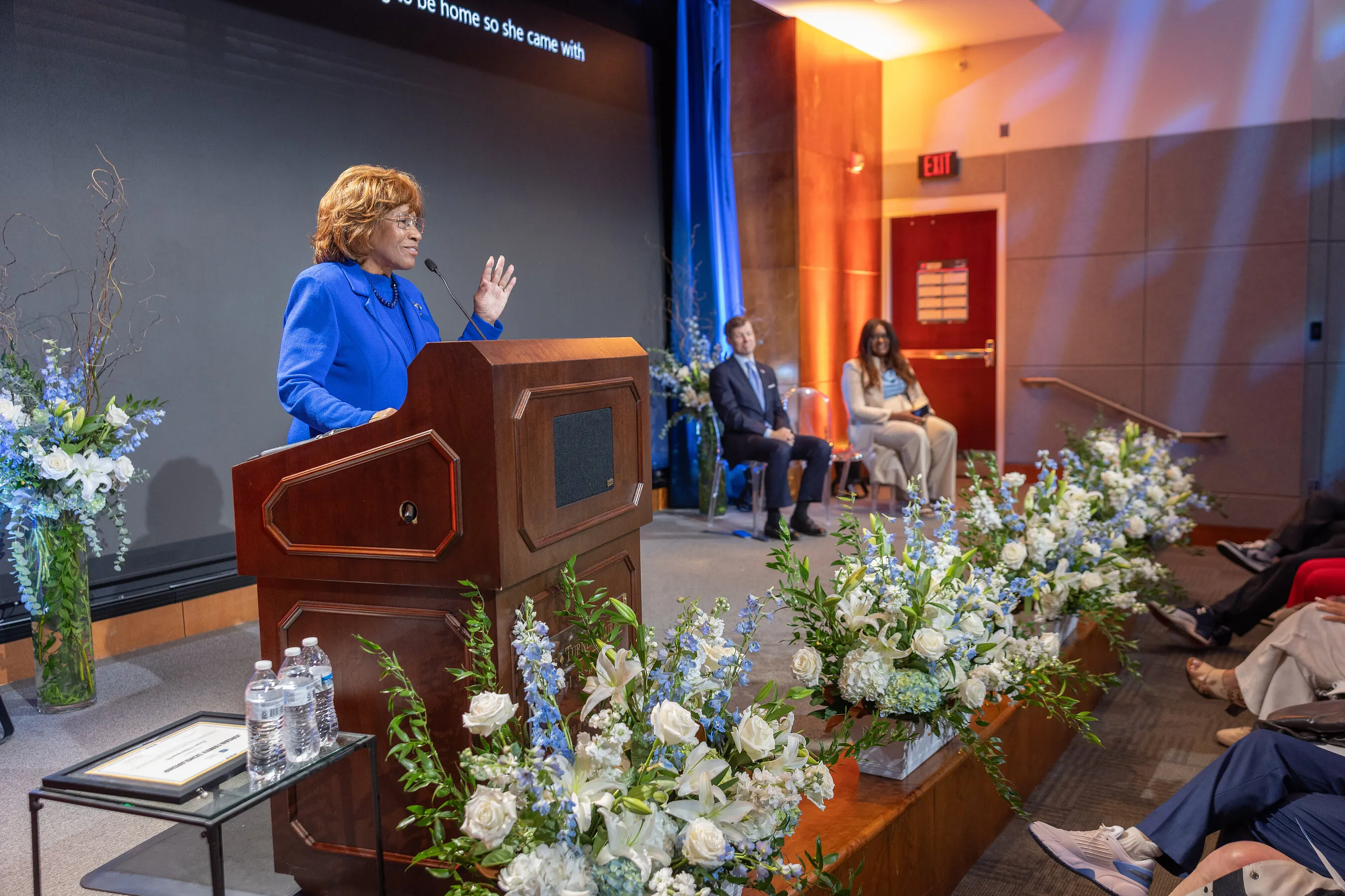 Deloris P. Jordan speaking at a podium on a flower-lined stage.