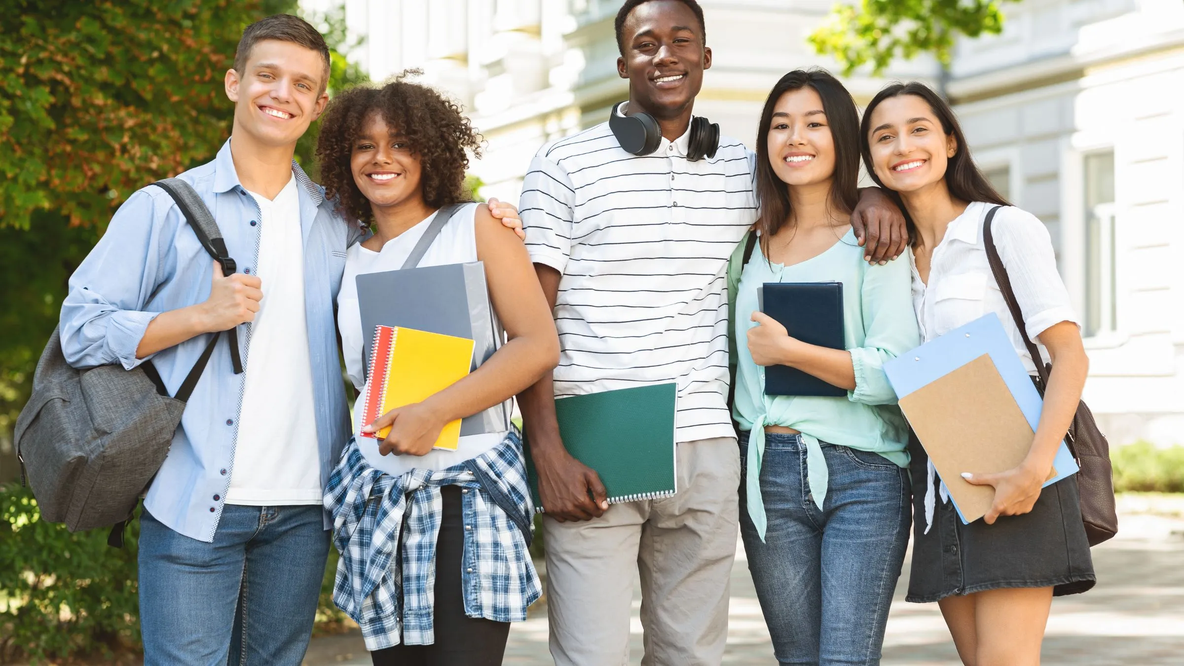 A group of smiling students holding books and folders outside a university building