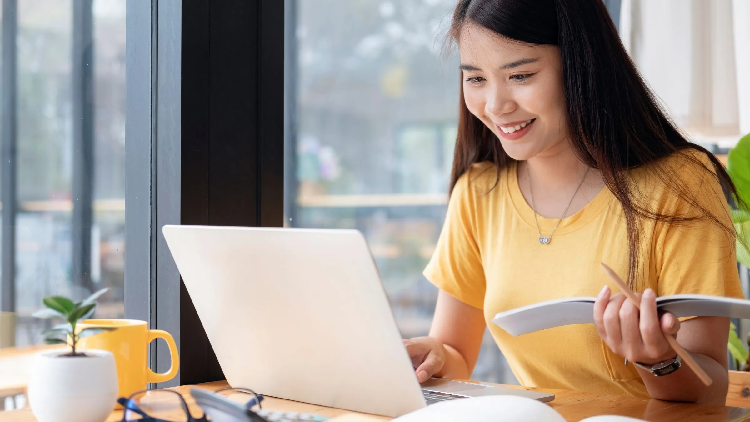 A student smiling at her laptop while holding an open book at a sunny desk