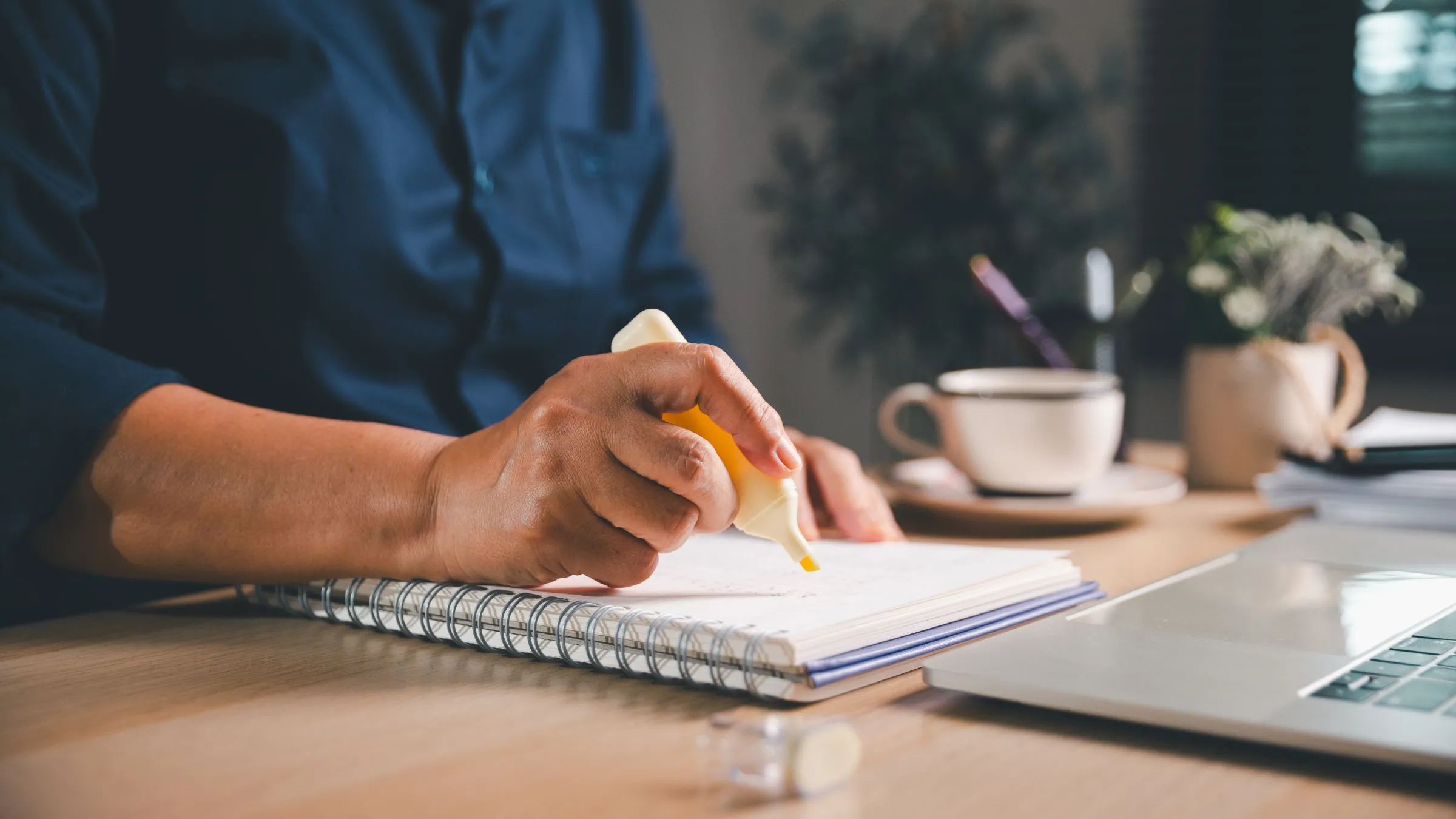 A hand marking notes in a spiral notebook with a highlighter beside a laptop
