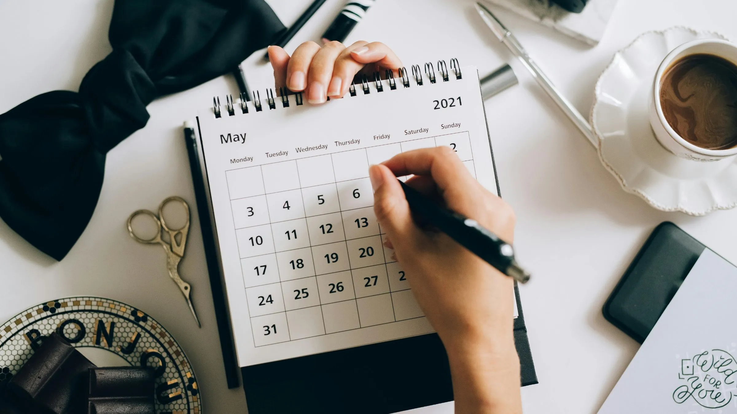 A hand writing dates on a desk calendar beside a cup of coffee
