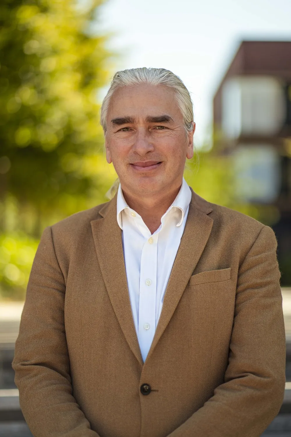 Dr Kevan Manwaring, gray-haired, in a tan blazer and open-necked white shirt, standing outdoors with trees and a building behind him.