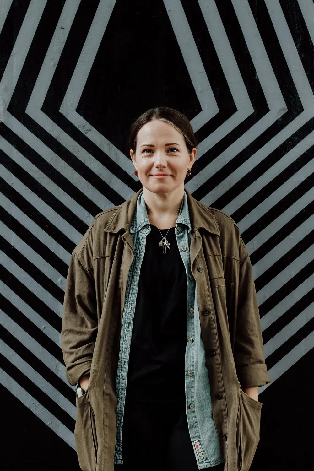 Louisa Flynn in an olive jacket over a black top, standing in front of a black and white mural of nested chevron stripes.