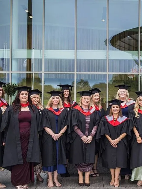 A group of graduates in caps and gowns standing outside a glass-fronted building.