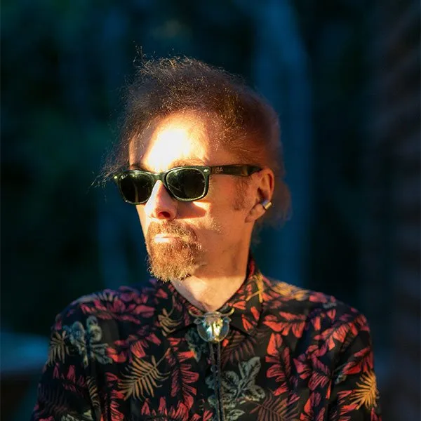 T.C. Boyle in dark sunglasses and a patterned shirt with a bolo tie, lit by low evening sun against a dark background.
